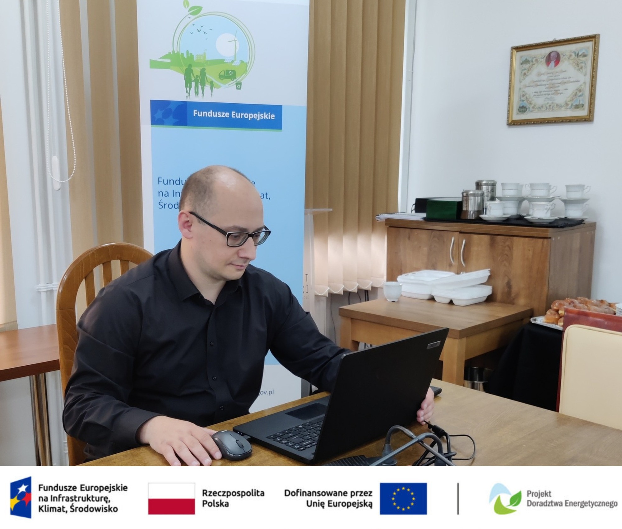 Zdjęcie przedstawia Doradcę Energetycznego z Wojewódzkiego Funduszu Ochrony Środowiska i Gospodarki Wodnej w Krakowie, siedzącego przy biurku i prowadzącego webinarium online. W tle widać roll'u Funduszy Europejskich